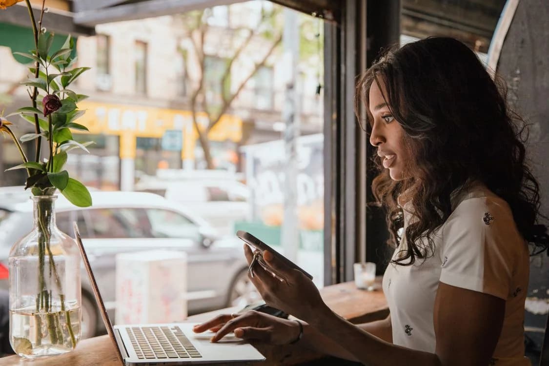 Mulher a trabalhar no computador e a consultar o celular junto à janela de um café.